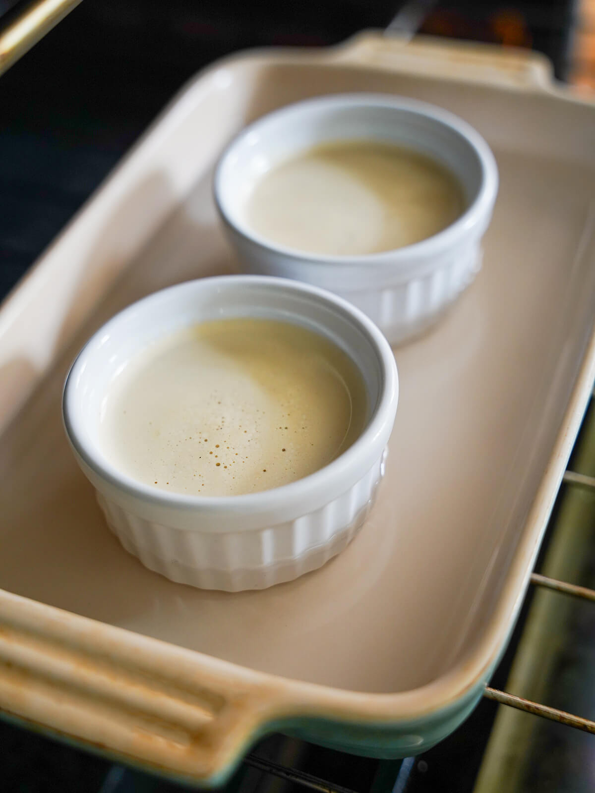 dishes filled with custard mixture in baking dish with water sitting on oven shelf.