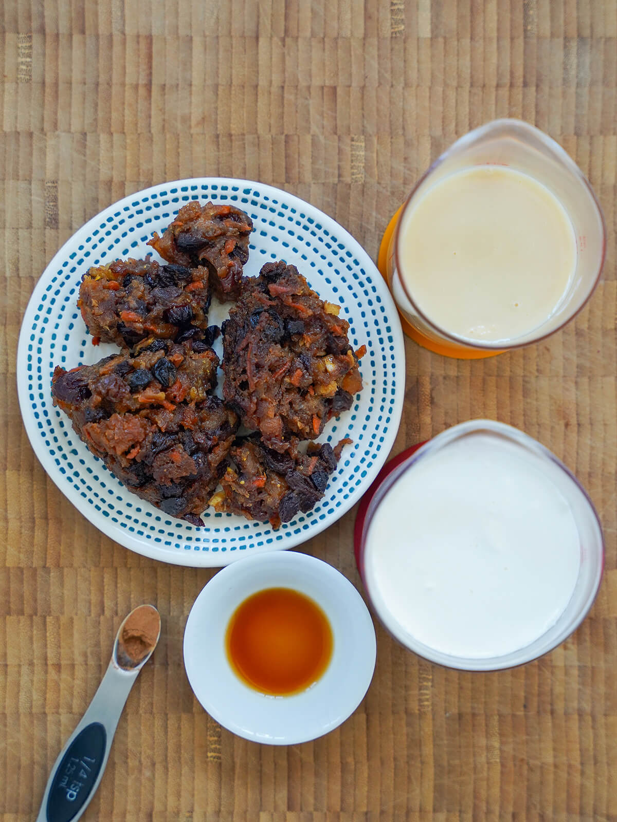 Christmas pudding chunks on plate with measures of cream and condensed milk to side, small dish with vanilla and cinnamon in scoop.