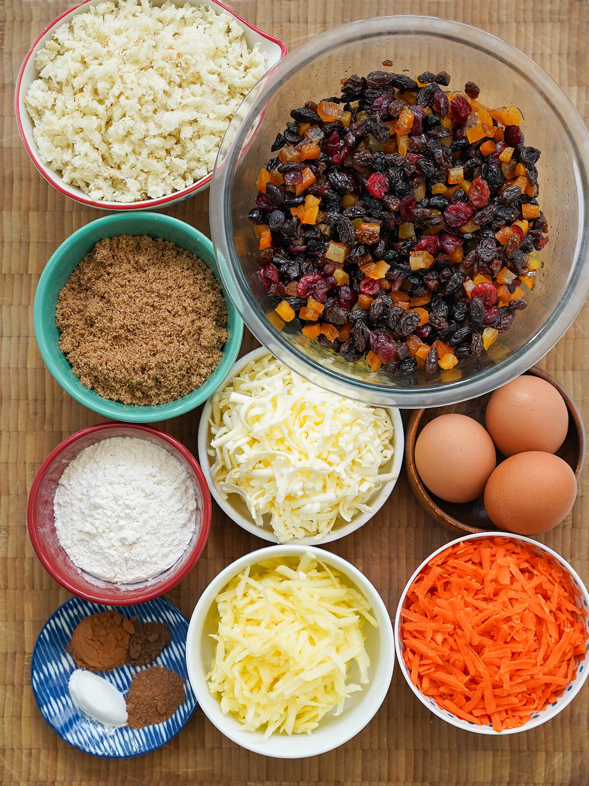 bowl of mixed dried fruit with bowls of grated carrot, butter, apple, breadcrumbs, sugar, flour, egg and spices around.