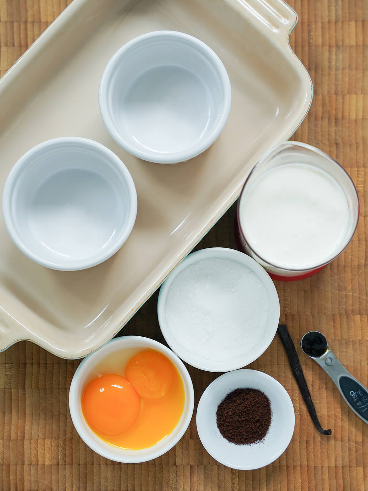 ramekins sitting in a baking dish with a measure of cream, bowl of sugar and bowl of egg yolks next to it with small dish with espresso powder and vanilla pod.