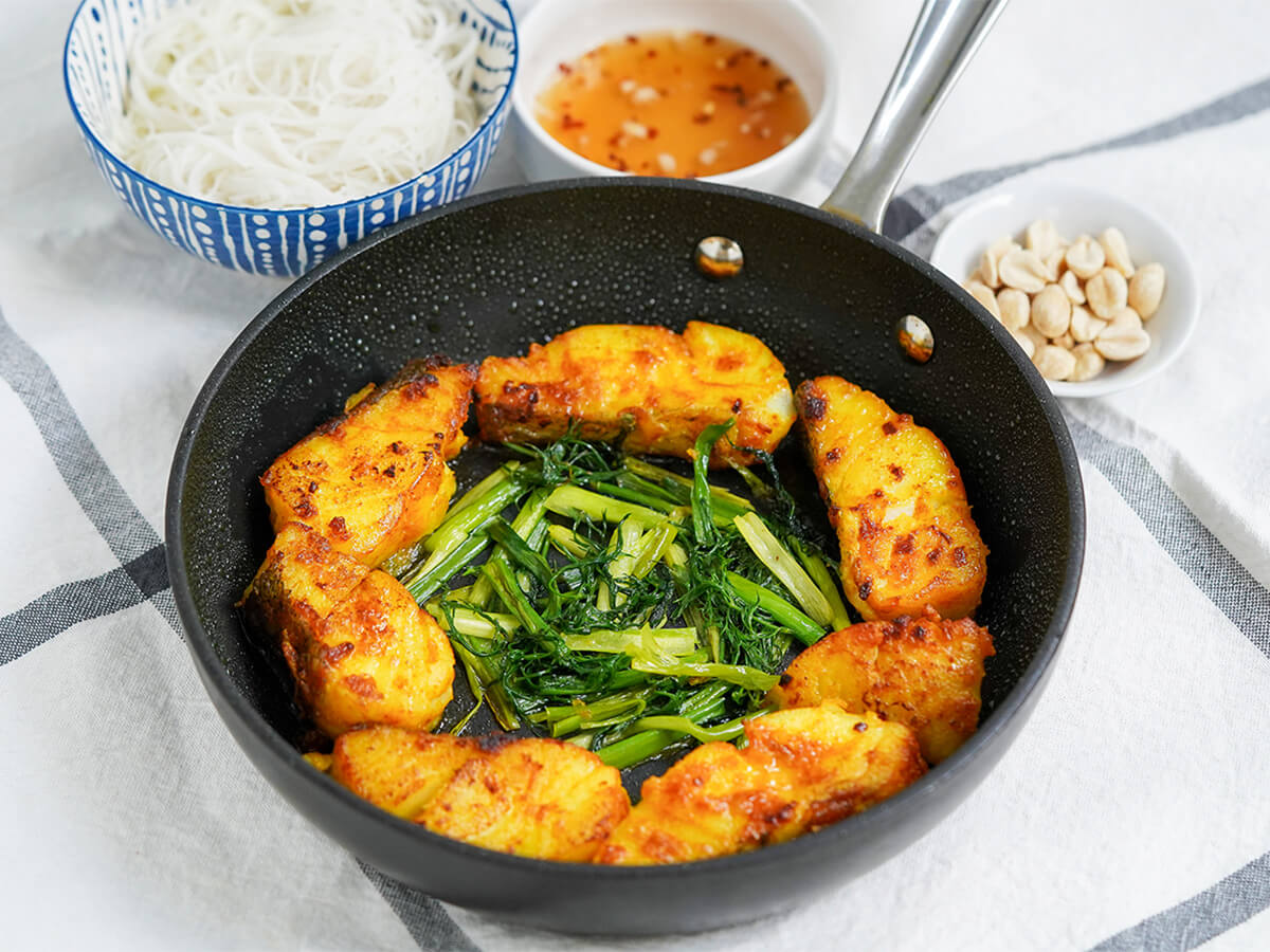 skillet of Vietnamese turmeric fish, cha ca la vong from side with noodles, sauce and peanuts behind.