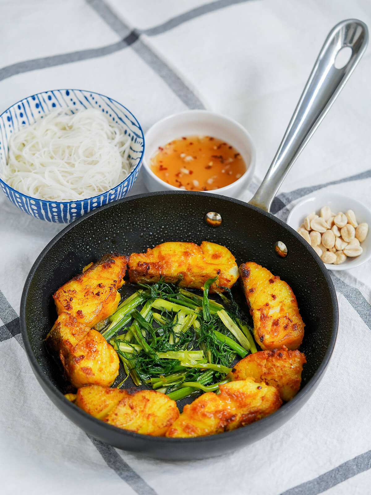 skillet of Vietnamese turmeric fish, cha ca la vong, with bowls of rice vermicelli noodles, dish of sauce and peanuts behind.