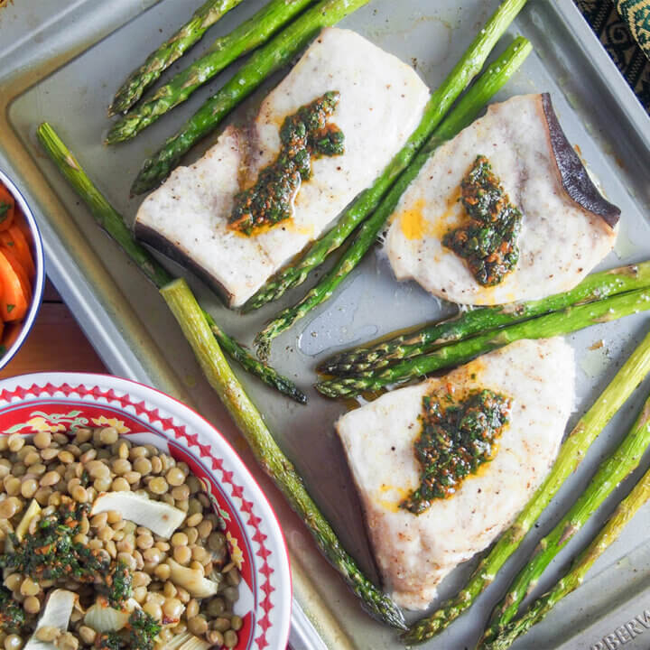 baked swordfish and asparagus on baking sheet with chermoula on top of fish and bowl of lentils in corner.