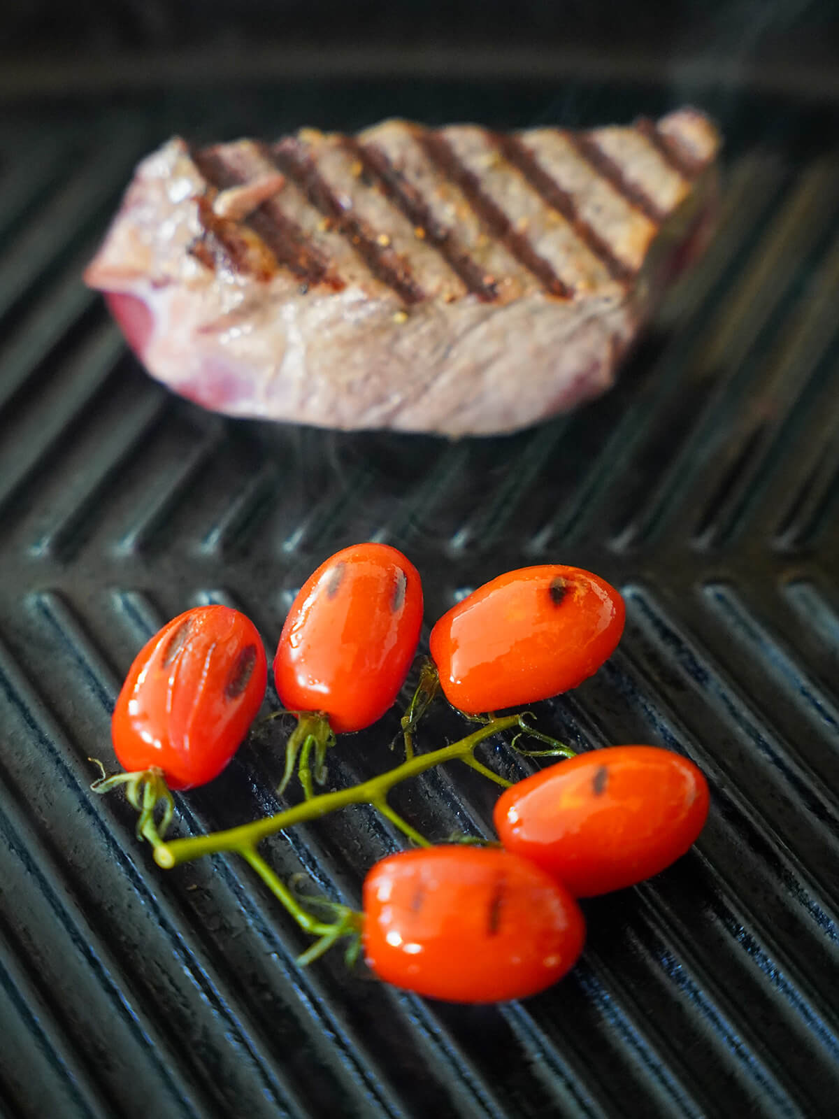 grilling a piece of steak and a small bunch of tomatoes on grill pan.