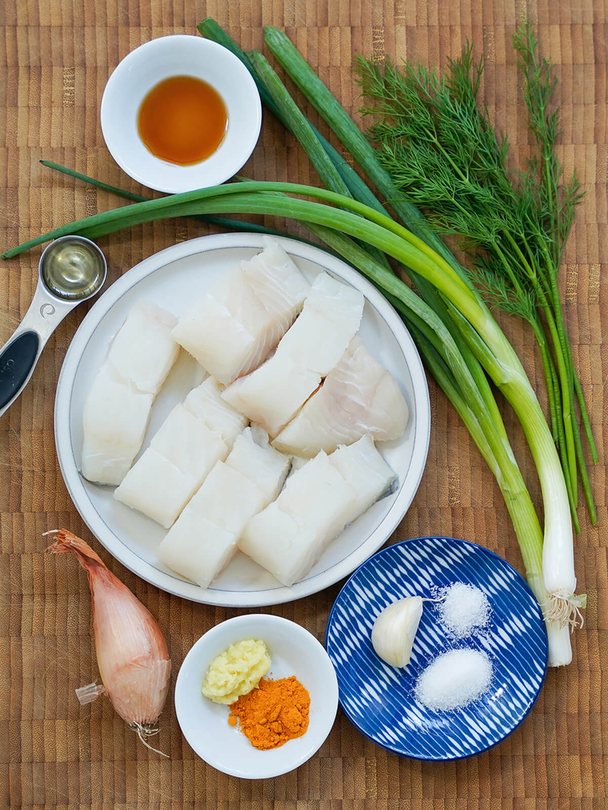 plate with pieces of fish with scallions and dill to side, dishes with garlic, turmeric, crushed ginger below and shallot to siide.