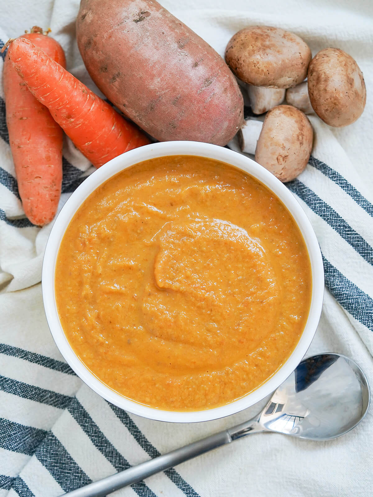 bowl of mushroom carrot and sweet potato soup with spoon below bowl and carrots, sweet potato and some mushrooms above.