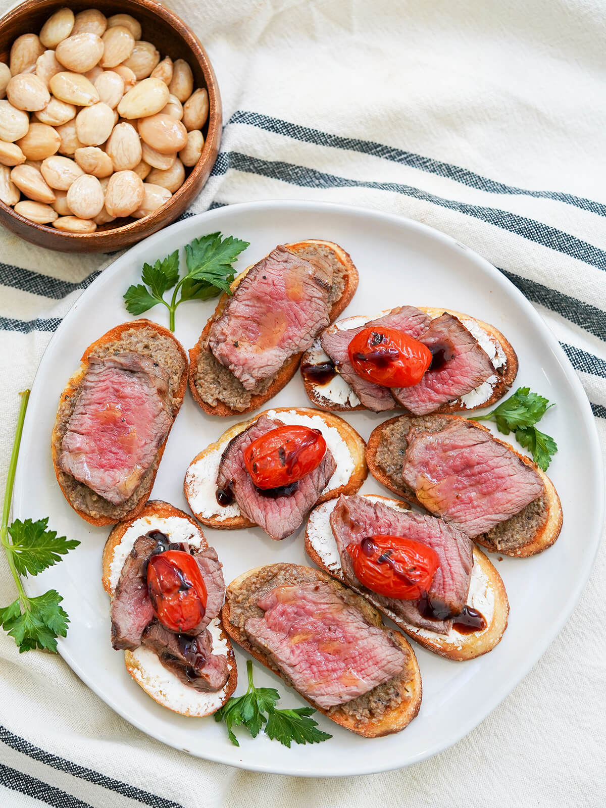 overhead view of plate of steak crostini, some topped with tomatoes, with bowl of nuts in corner.