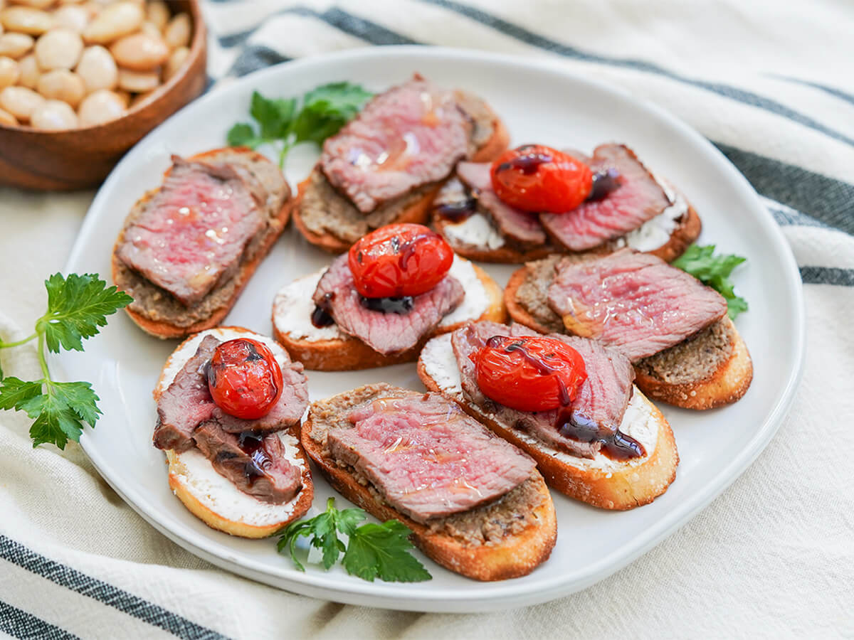 plate of steak crostini, some with pate under steak and others with goat cheese under and tomato and balsamic on top.