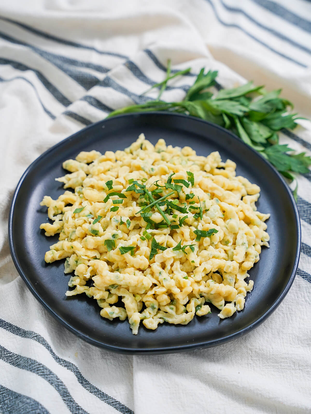 Herb spaetzle are easy to make and tasty both as a side or on their own with butter, herbs and optional cheese. Deliciously comforting. plate of herb spaetzle with herbs on top and more herbs behind plate.