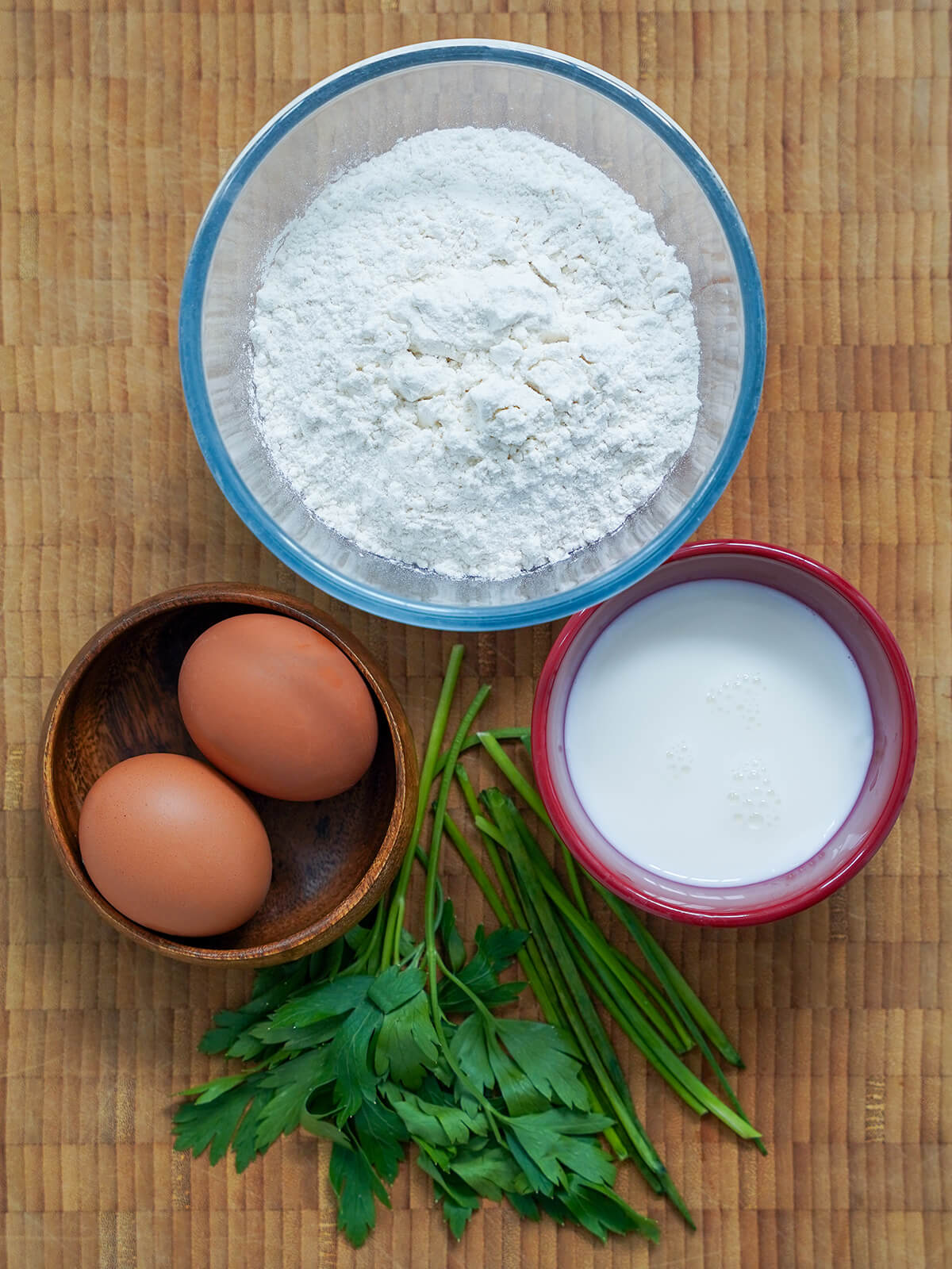ingredients for herb spaetzle bowl of flour, smaller dishes of milk and with eggs in, chives and parsley stems between bowls.