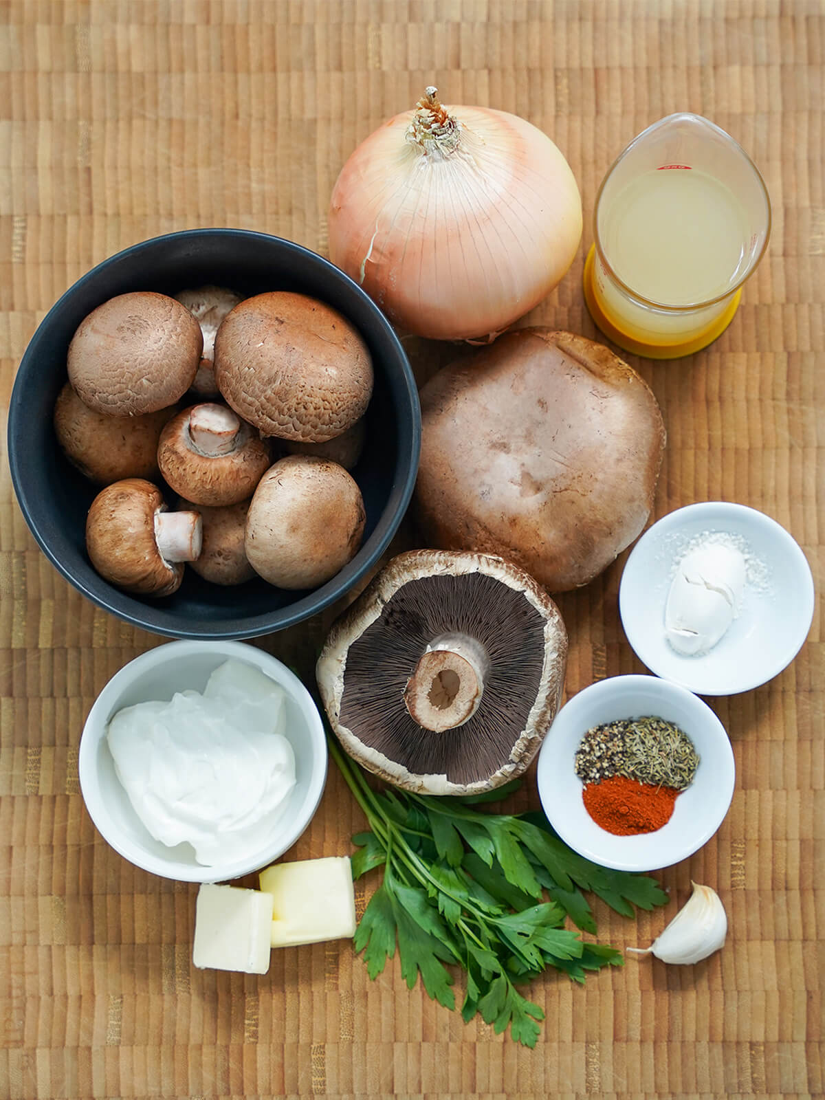 ingredients for mushroom stroganoff bowl of mushrooms, onion, some portobello mushrooms, parsley, dish with sour cream, measure of stock, chunks of butter and small dishes with flour and spices all on chopping board.