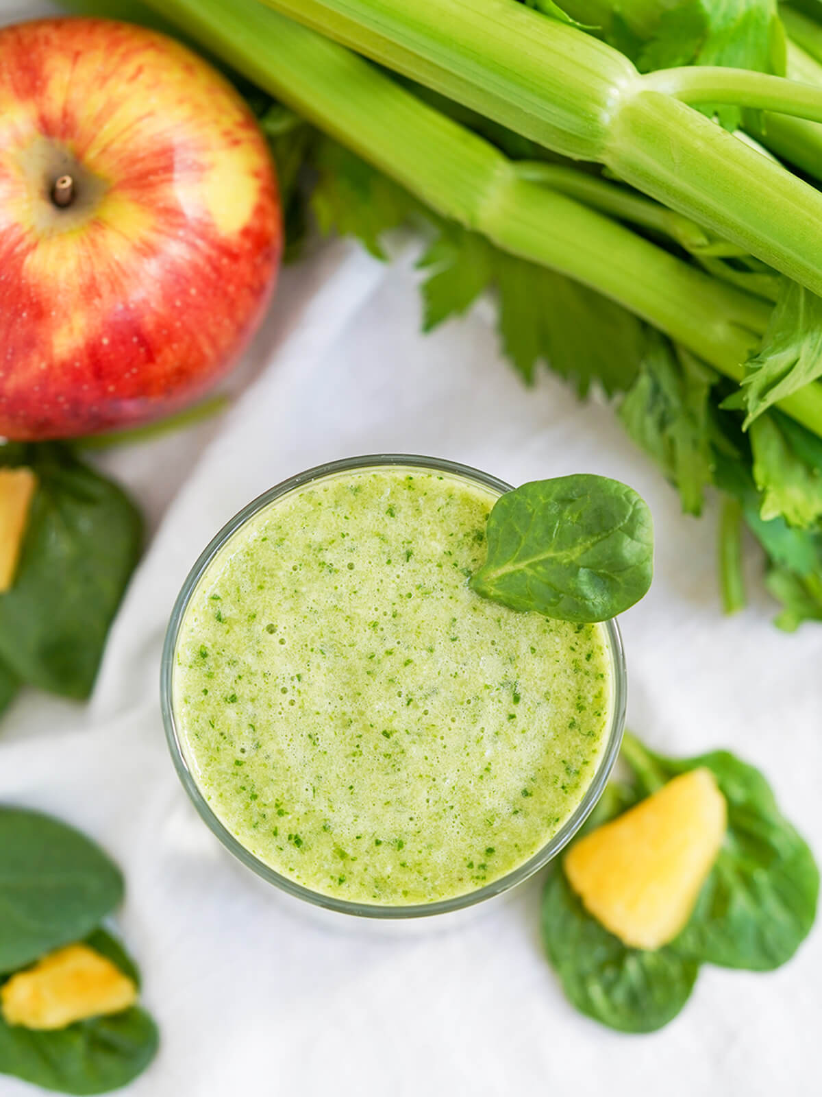 overhead view of a glass of pineapple and spinach smoothie with bunch of celery and apple in top corners.