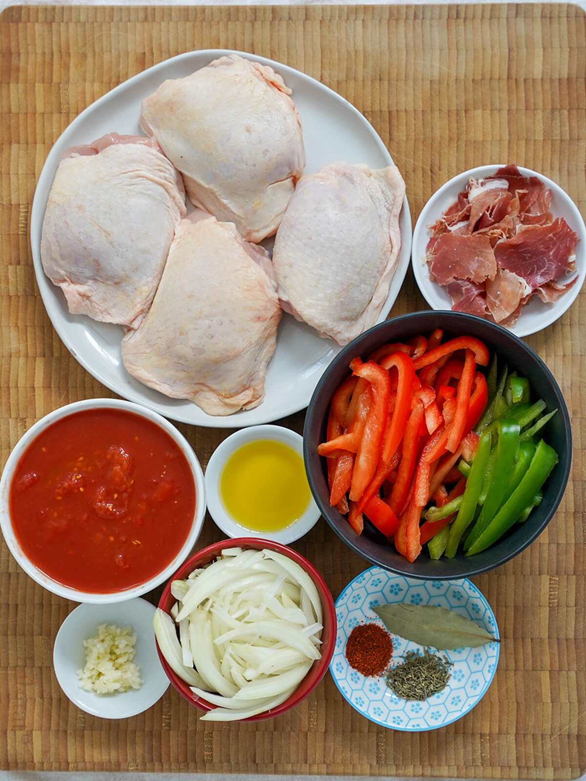 chicken thighs on a plate, bowls with sliced peppers, onion, chopped tomatoes, ham, garlic and herbs.