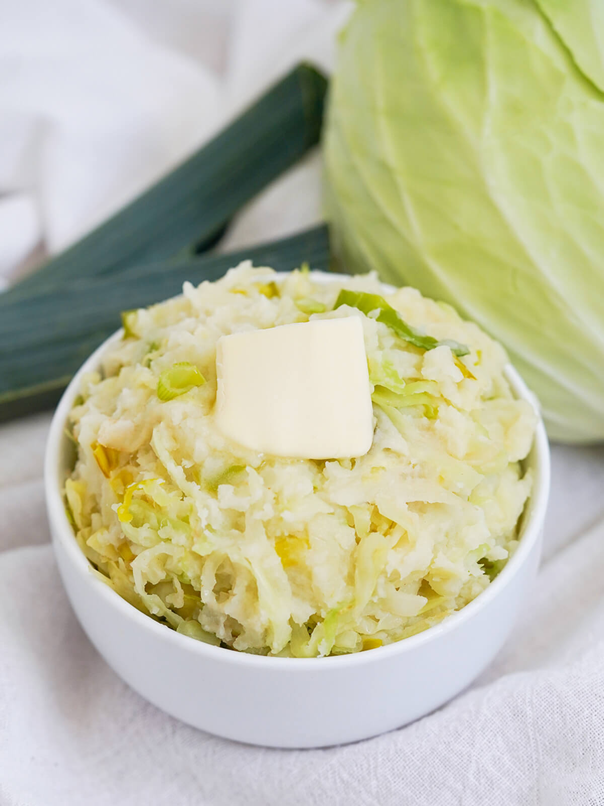 bowl of colcannon, mashed potatoes with cabbage, topped with butter with cabbage and leek top in background behind.