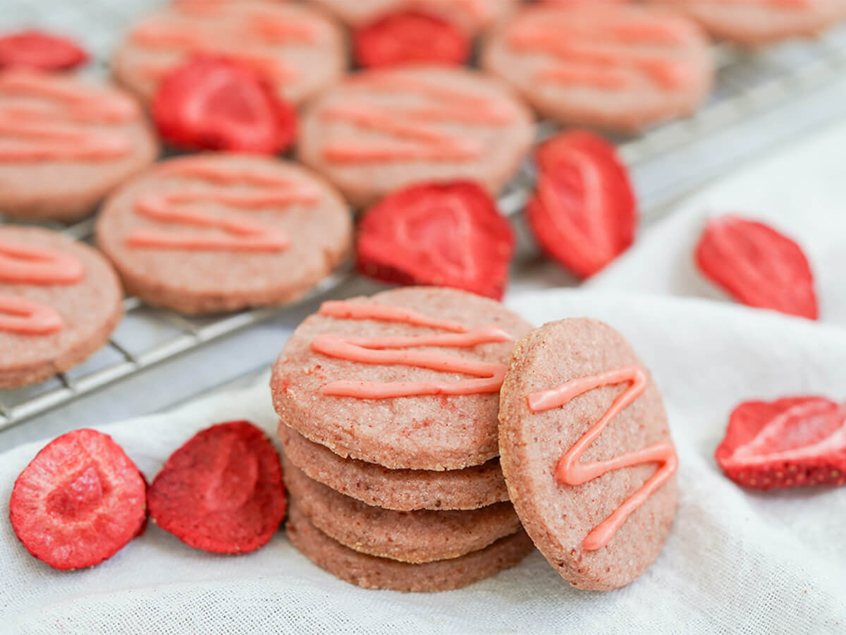 small stack of strawberry shortbread cookies with freeze dried strawberry pieces around and more cookies behind.