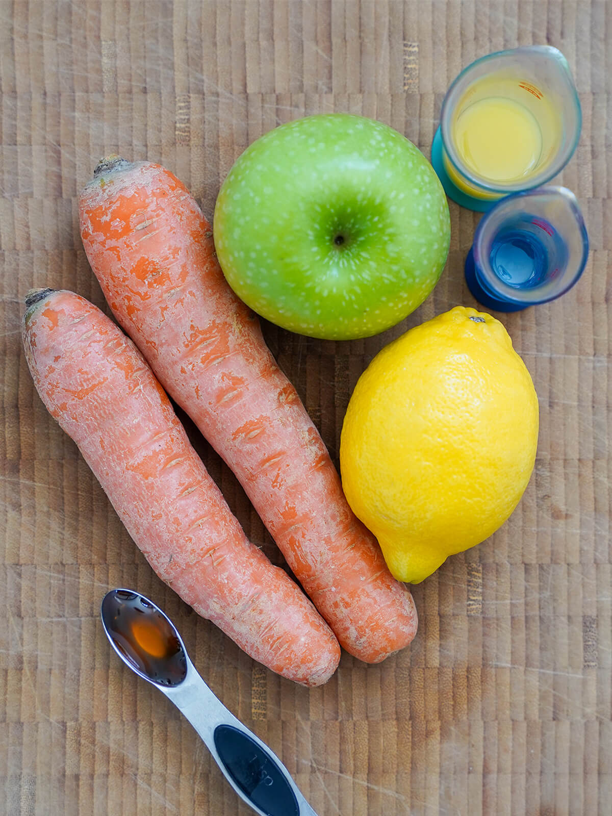 two carrots, a green apple, lemon on board with measures with honey, orange juice and oil to sides.