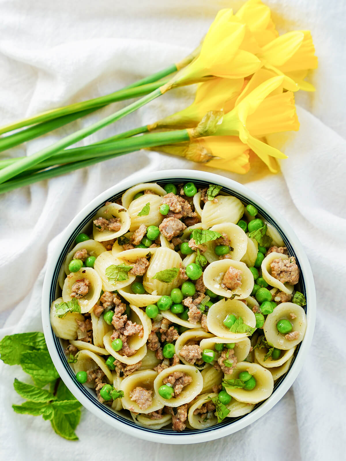 This pea and lamb pasta is easy to make and packed with tasty spring flavors, with the rich lamb balanced out by fresh mint, peas and a touch of spice. Quick, tasty and a great easy meal. overhead view of a bowl of pea and lamb pasta with daffodils above bowl.