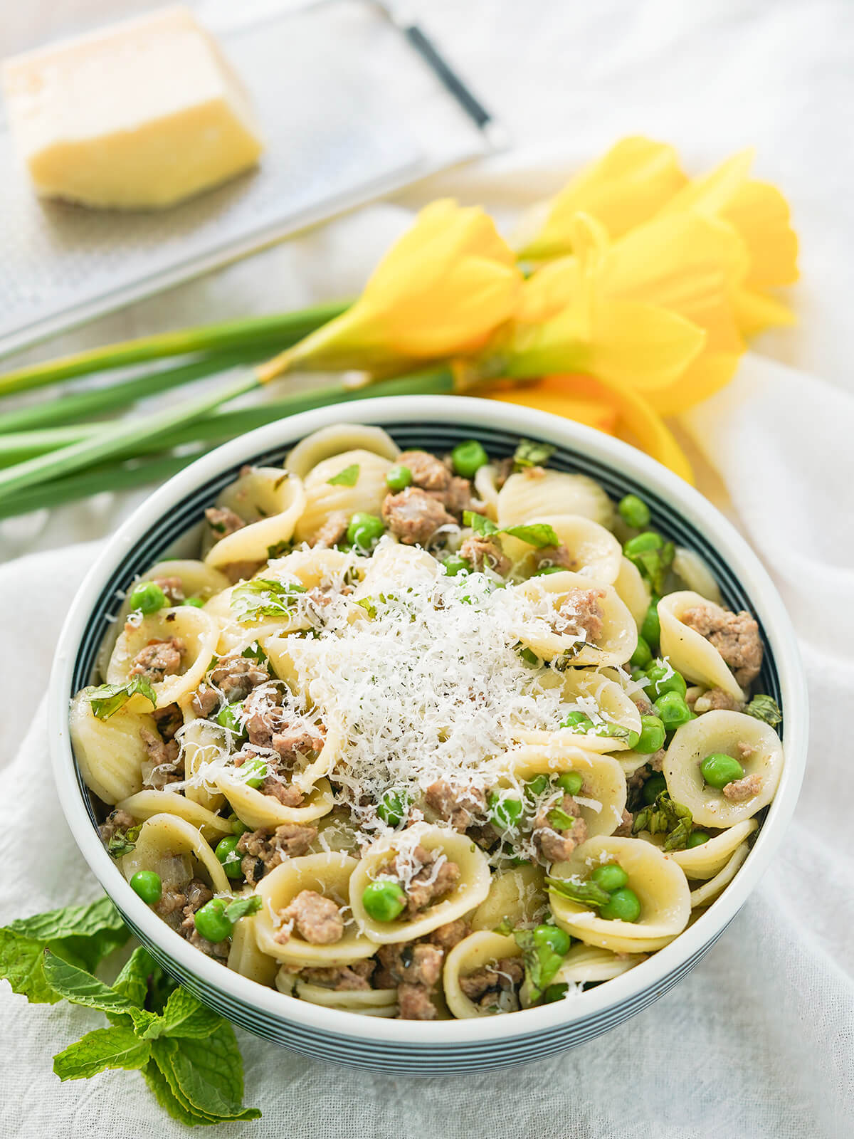 This pea and lamb pasta is easy to make and packed with tasty spring flavors, with the rich lamb balanced out by fresh mint, peas and a touch of spice. Quick, tasty and a great easy meal. bowl of pea and lamb pasta with bunch of daffodils and grater with cheese in background.