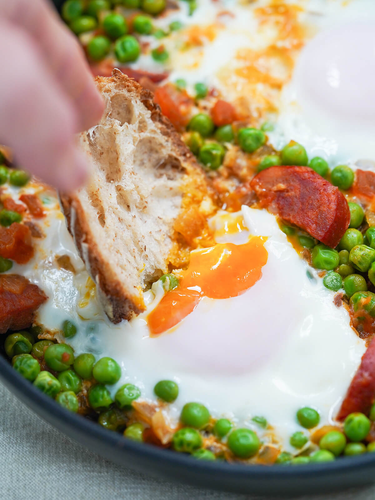 dipping a piece of bread into egg yolk with peas and chorizo around it in skillet.