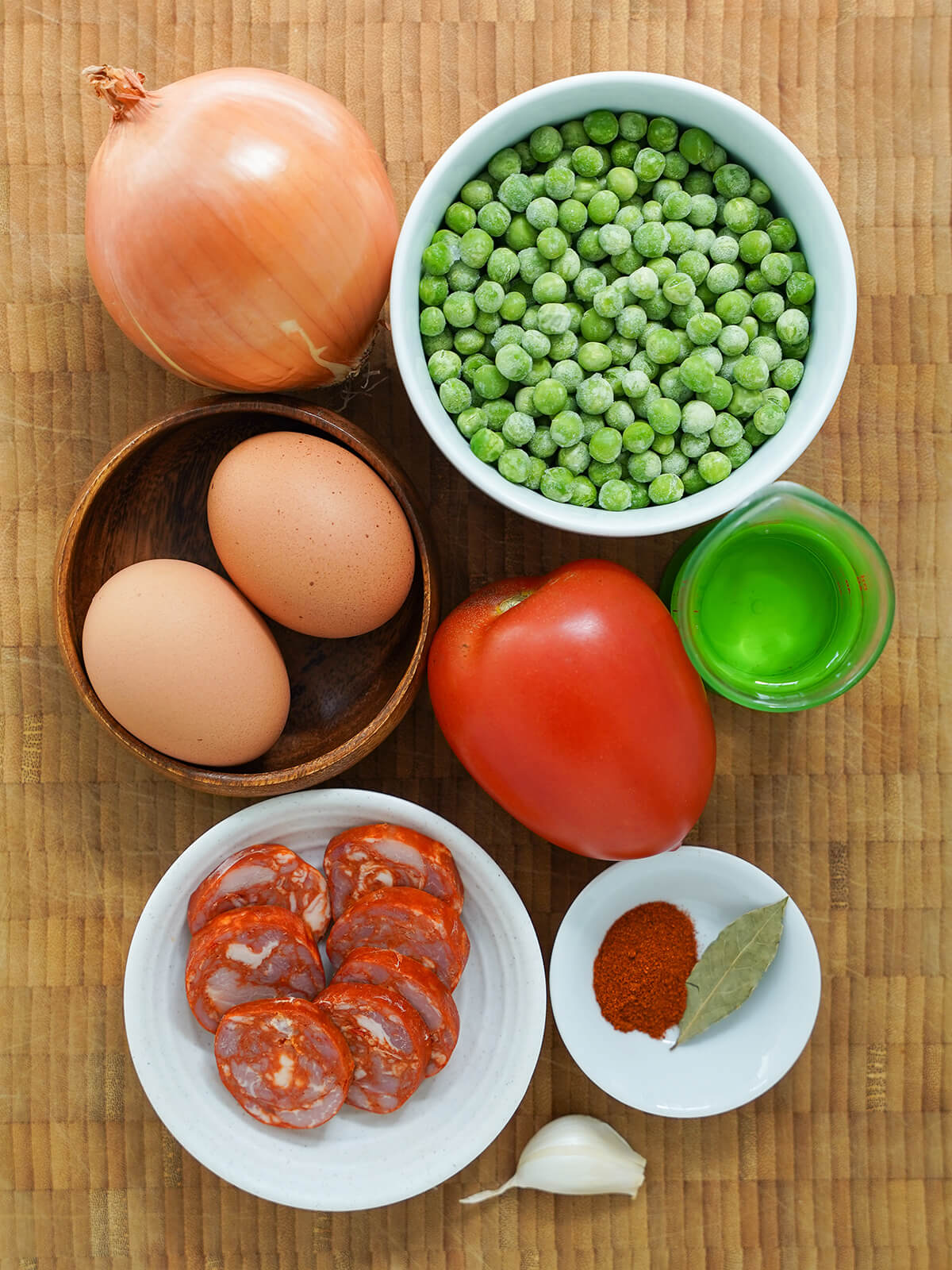 bowl of peas, eggs, tomato, sliced chourico, an onion, measure of wine and garlic, bay leaf and paprika in dish all on wooden board.
