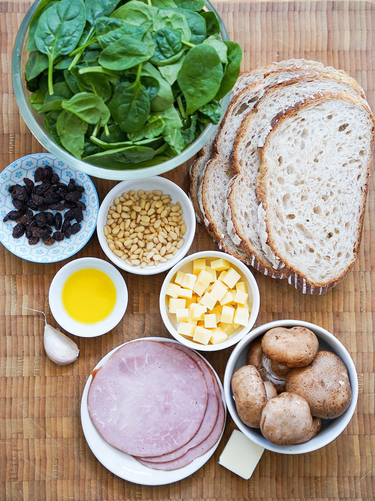 bowl of spinach, small dishes with raisins, pine nuts, cubes of cheese, mushrooms and olive oil with stack of sliced bread to one side and garlic to other.
