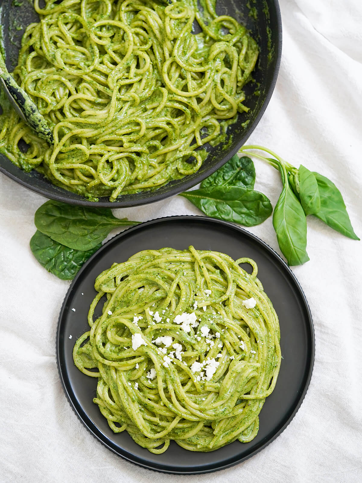 Tallarines verdes is Peruvian pesto that's that bit different from the classic, but no less tasty. Easy, creamy, smooth and comforting, it's great for busy nights or any excuse. overhead view of plate of tallarines verdes, also called Peruvian pesto, with skillet with more in it above and spinach and basil leaves in between.