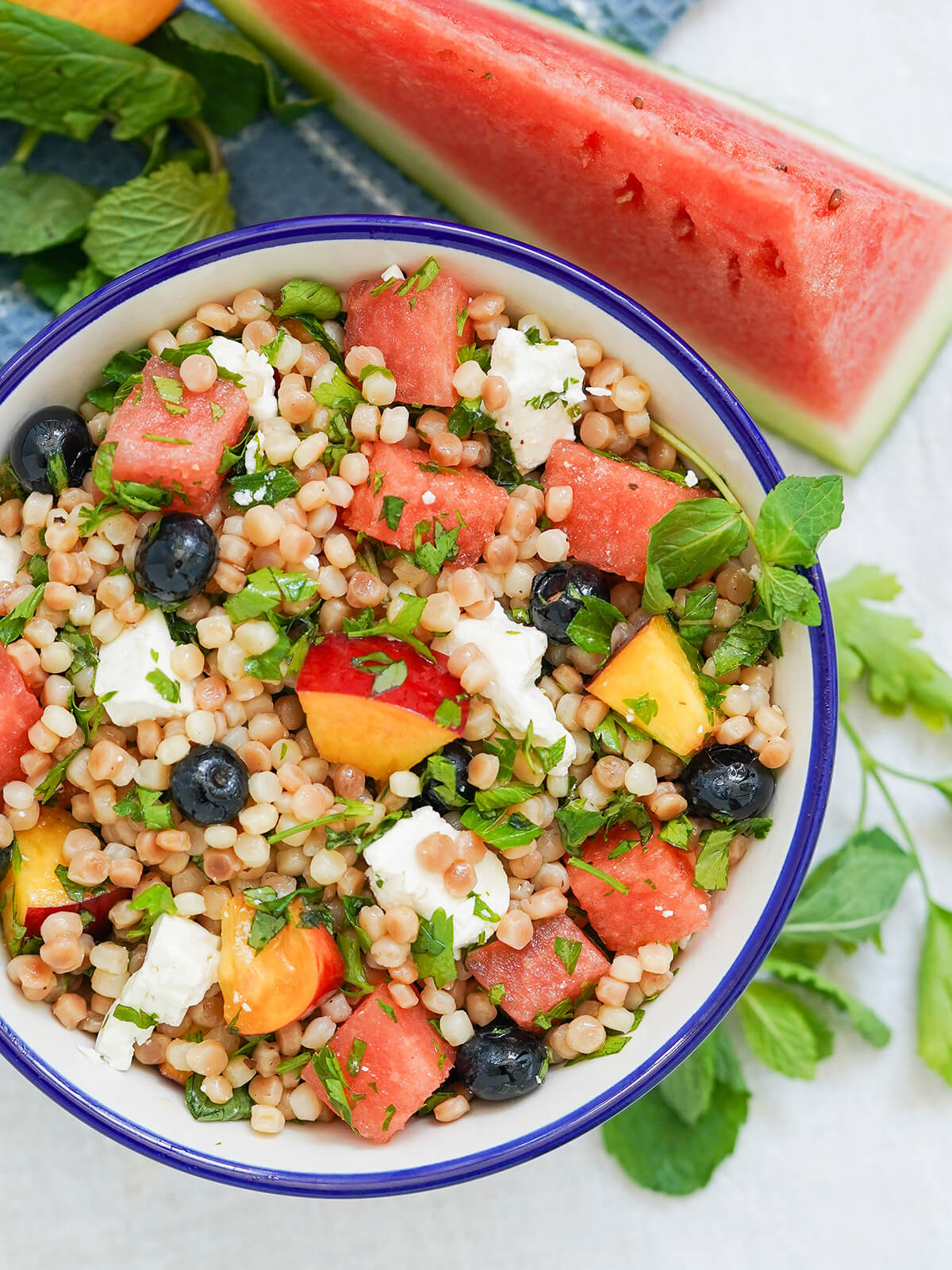 overhead part view of bowl of fruit couscous salad with herbs around bowl and wedge of watermelon above.