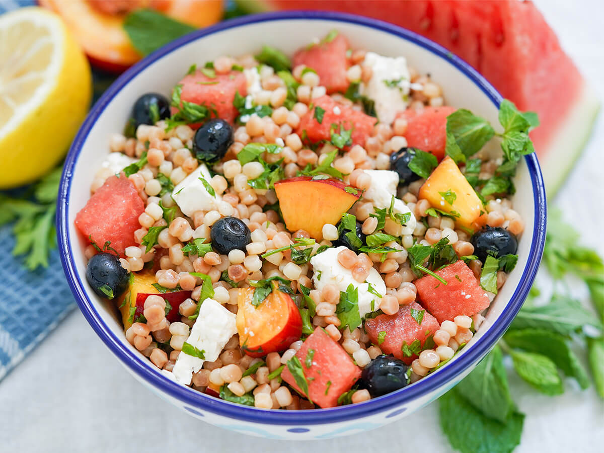 bowl of fruit couscous salad with herbs around and watermelon, lemon and nectarine blurred behind.