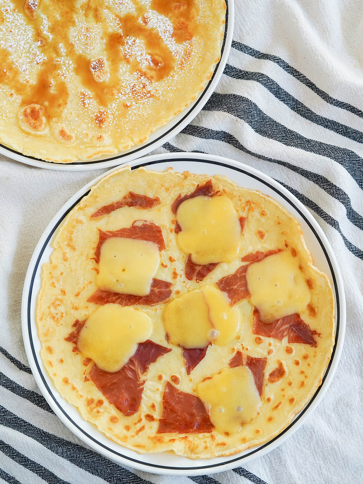 plates of sweet and savory pannekoeken, Dutch pancakes overhead view of two plates of Pannekoeken Dutch pancakes with one topped with ham and cheese and other syrup and powdered sugar.