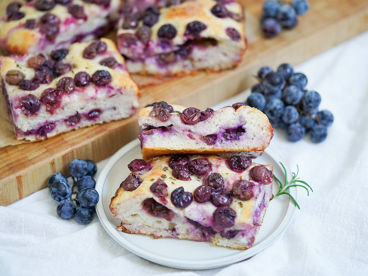 slices of Schiacciata all’uva grape focaccia on plate with grapes wither side and more slices on board behind.