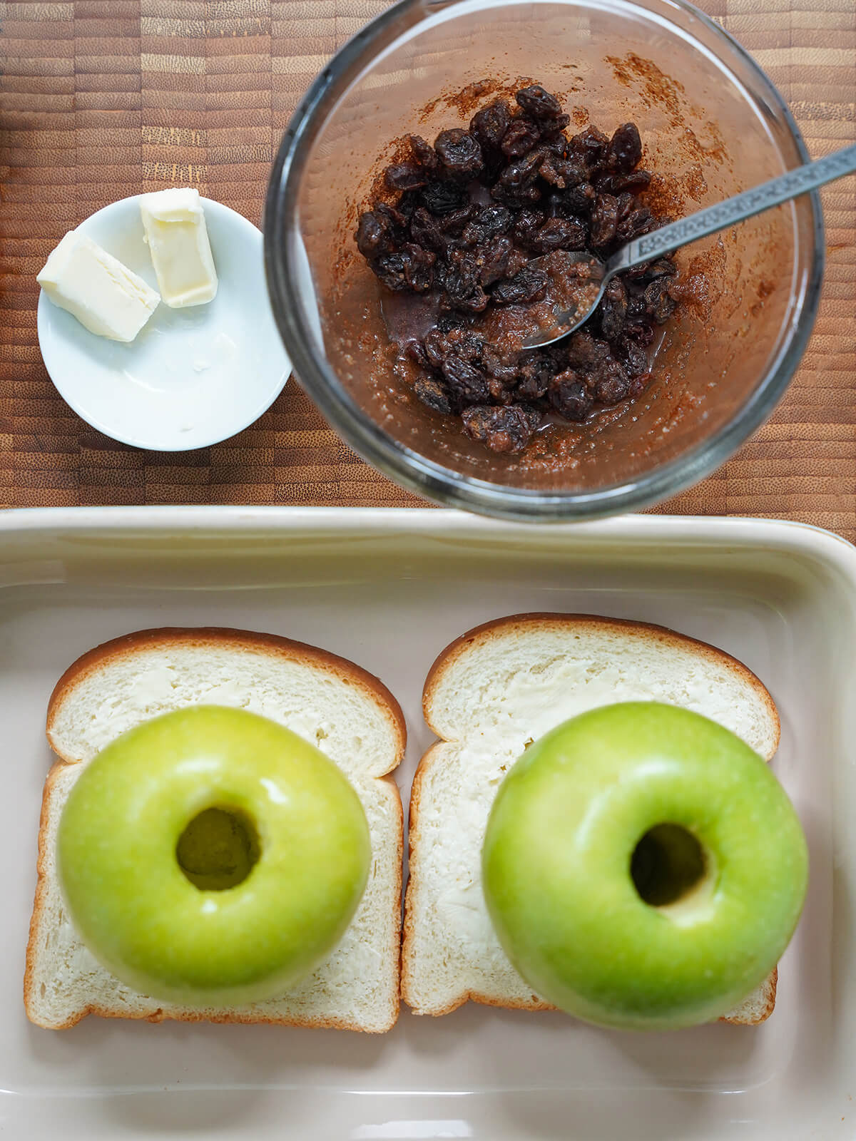 bowl of raisin mixture, pieces of butter above and cored apples on top of slices of bread below.