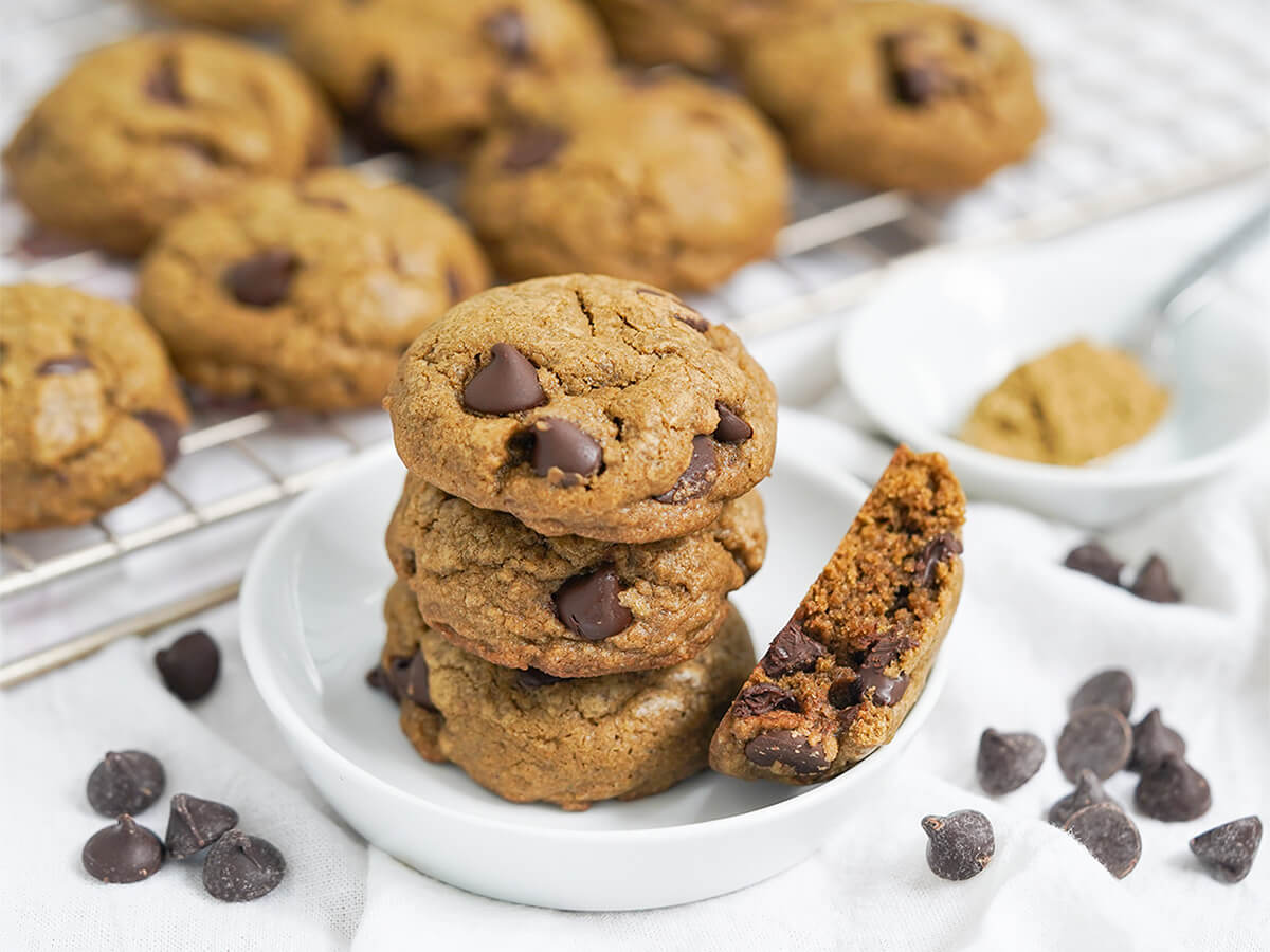 stack of hojicha chocolate chip cookies on small plate with more on cooling rack behind and chocolate chips either side.