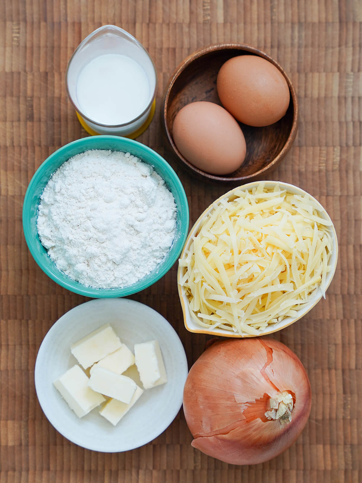 bowls with eggs, flour, grated cheese, butter, measure of milk and onion below on wooden board.