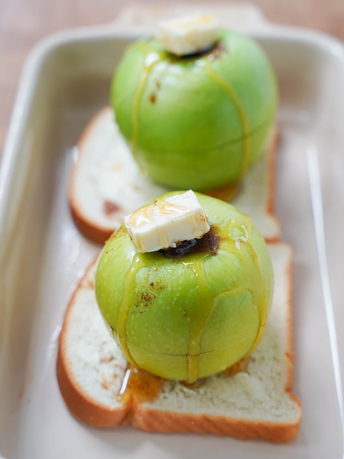 stuffed apples topped with butter and drizzle of honey on top of slices of bread in dish before baking.