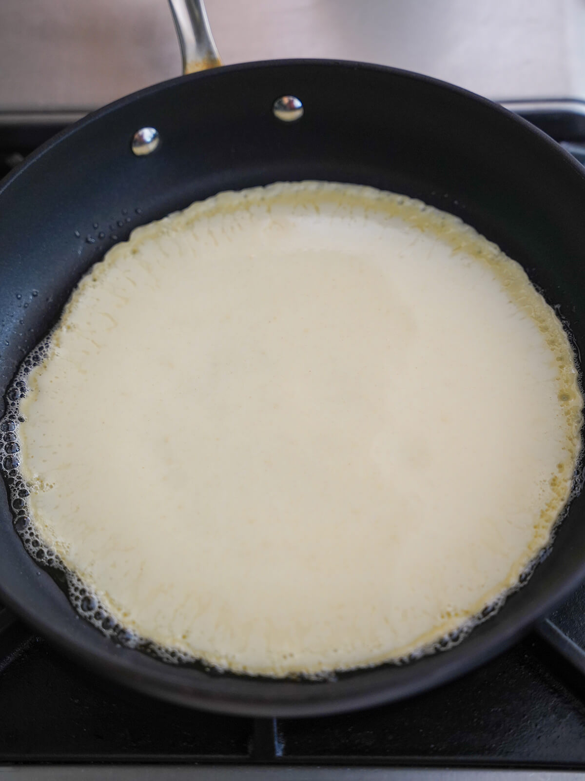 starting to cook Pannekoeken Dutch pancakes pancake batter in skillet with bubbles of butter around edges.